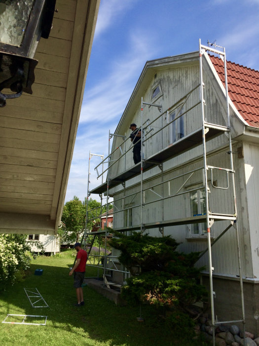 Två personer arbetar på byggnadsställning vid vit träfasad på hus under underhållsmålning.