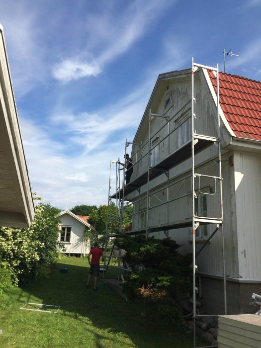 Två personer reser en byggnadsställning vid en vitmålad husfasad under en solig himmel.