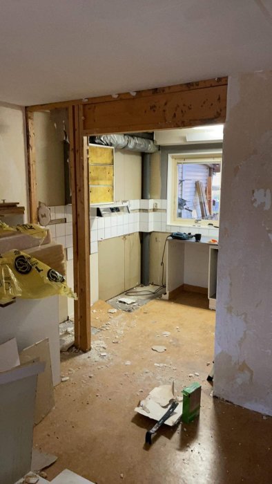 Byggeområde med delvis fjernet vegg til kjøkkenet, eksponert bjelke, rotete gulv og verktøy i et rom under renovering.