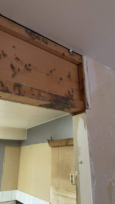 Revne vegger ved kjøkkeninngang, delvis avdekket bjelke over åpningen. Veggene har synlig slitt gips og en stikkontakt ved siden av.