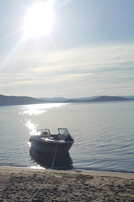 Motorbåt förtöjd vid strandkant med solnedgång och glittrande vatten i bakgrunden.