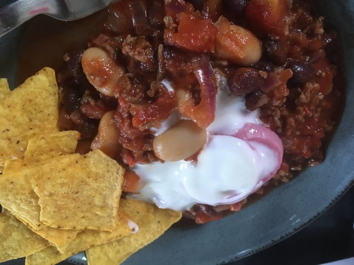 Chili con carne med grekisk yoghurt, picklad rödlök och nachos, serverad i en skål.