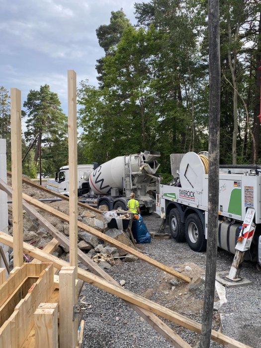 En byggarbetsplats där en person övervakar gjutning av betong från en cementbil med skog i bakgrunden.