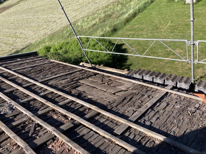 Avskalat tak med träbrädor, några staplade betongpannor och byggställning vid takfot under renoveringsarbete.