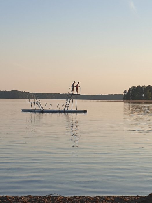 Två personer står på en brygga med dyktorn på en lugn sjö vid skymningen.