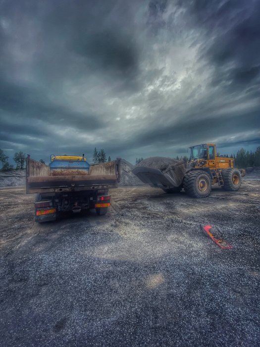 Lastbil och hjullastare på grustäkt under mulet himmel, lassar grus.