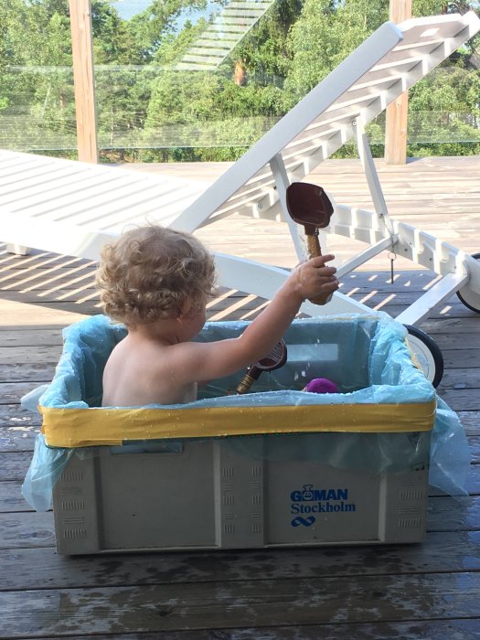 Ett barn leker i en improviserad pool gjord av en plastlåda på en träterrass.