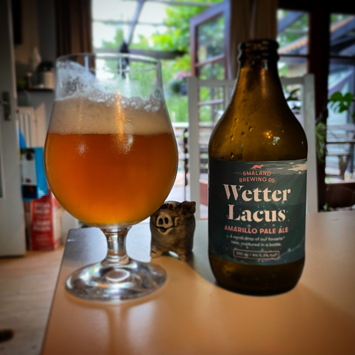 Ölglas halvfullt med öl bredvid en flaska Vetter Lacus Amarillo Pale Ale och en liten grå ugglefigur.