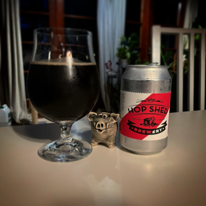 Ett mörkt ölglas bredvid en burk från Hop Shed Brewery och en liten grisfigur på ett bord.