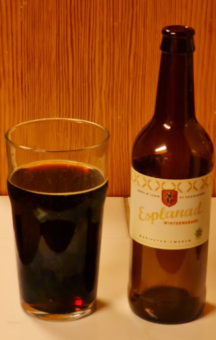 Ett glas mörkt öl bredvid en tom ölflaska med etiketten "Esplanad winter warmer" mot en träpanel.
