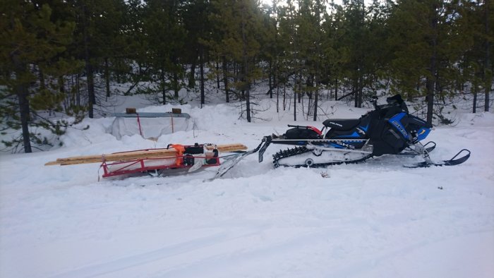 Blå arbetsbelastad snöskoter med släde och virke i snöig skogsmiljö.