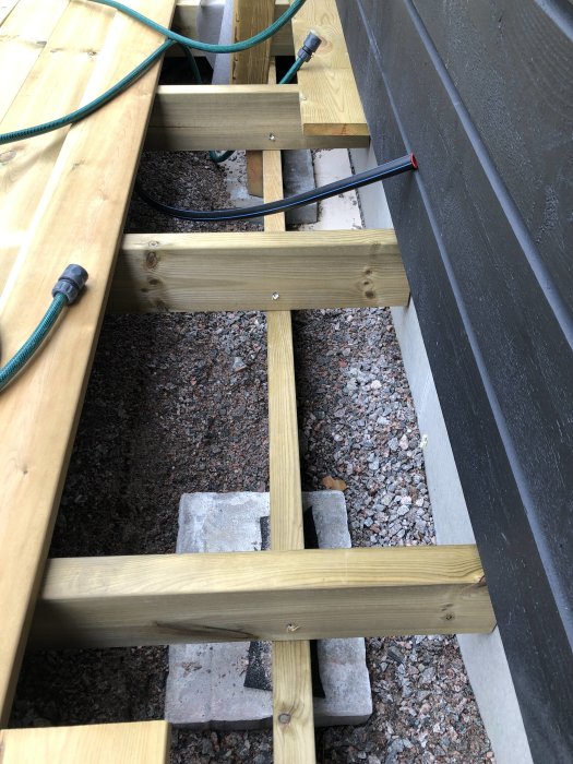 Konstruktion av en träaltan med plintar och 20x30 cm stenplattor längs husväggen, synliga dräneringsgrus och vattenledningar.