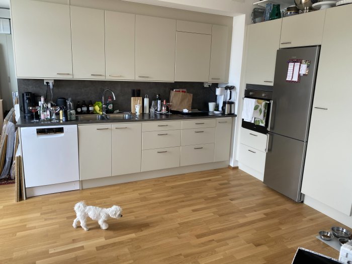 Kök före renovering med beige skåp och betongmönstrat stänkskydd, vit hund på golvet.