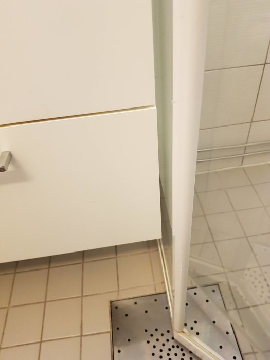 Smalt utrymme mellan en vit tvättställskommod och en duschkabin i ett badrum.