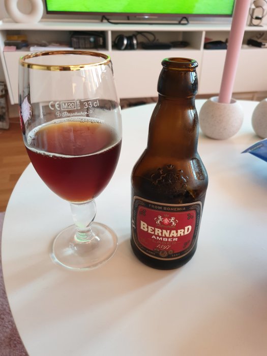 Glas med mörk öl bredvid en flaska Bernard Amber på ett vitt bord framför en TV som visar fotboll.