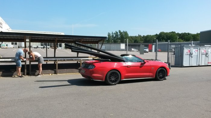 Röd cabriolet parkerad vid byggaffär med fyra avgasrör synliga, två personer i bakgrunden.