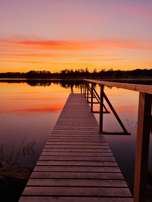 Träbrygga som sträcker sig ut i en stilla sjö under en färggrann solnedgångssky.