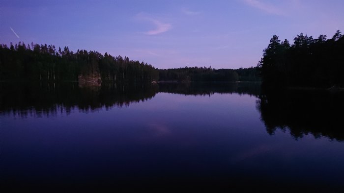 Skymningsvy över en stilla sjö med spegelblank yta och mörka träd längs stranden.