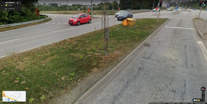 Trafikrondell i Lund med synlig ogräsbesvuxen konstgräsremsa, omgiven av asfalterade vägar och bilar i rörelse.