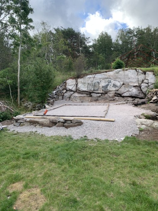 Grundläggning för växthus med markarbete och krossgrus på en gräsmatta, nära en stenbelagd slänt och träramar.