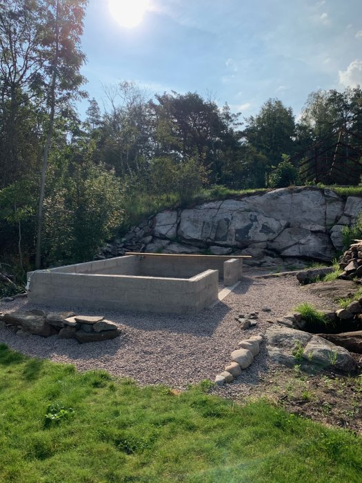 Nybyggt växthusfundament i en trädgård med grus, stenar och grönska under en solig himmel.