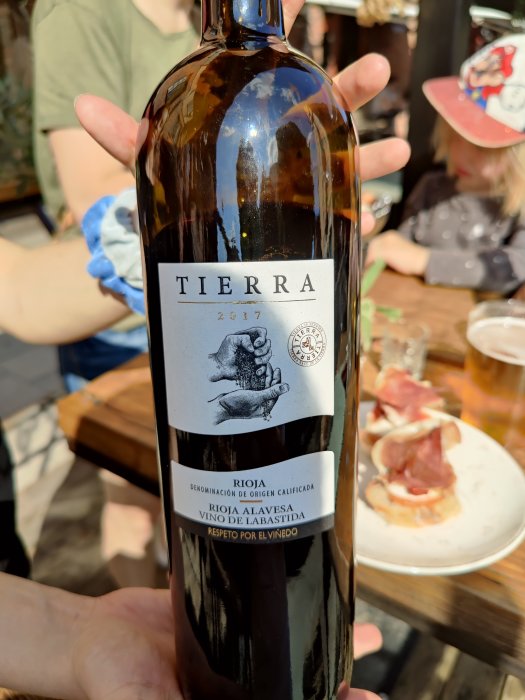 Hand som håller en flaska vin, TIERRA 2017 Rioja, med skinka på tapasrätter i bakgrunden.