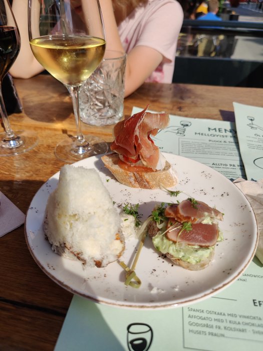 Ett glas vitt vin och en tallrik med tapas inklusive bröd med skinka och ost på en restaurangterrass i solsken.