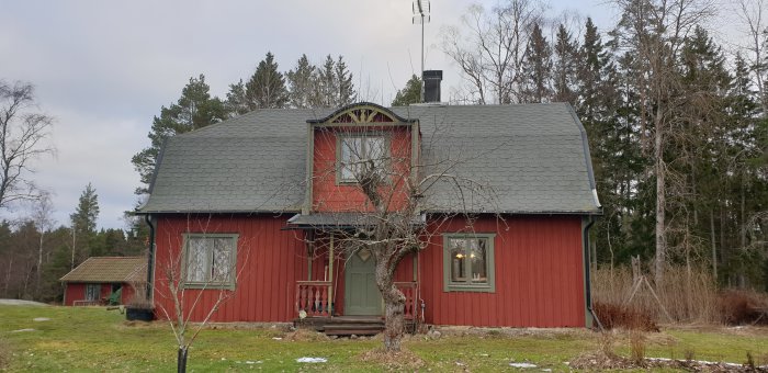 Ett traditionellt rött hus med grått tak och vit dekoration ovanför fönstret, delvis omgivet av träd och med lite snö på marken.