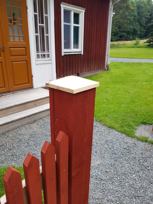 Ommålad röd grindstolpe med ny tillverkad stolphatt av trä framför ett rött hus med vita fönster.