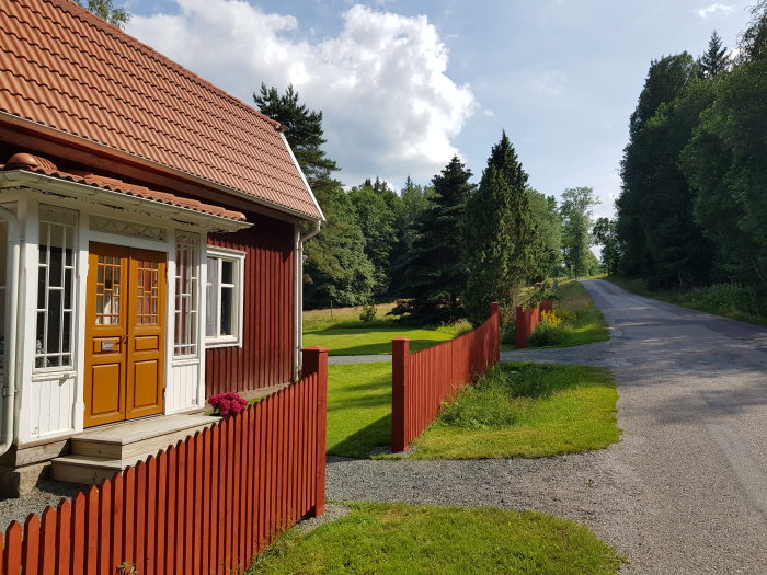 Rödmålat trästaket med nyinstallerade stolphattar vid en landsväg, intill en röd stuga med gult dörrparti.