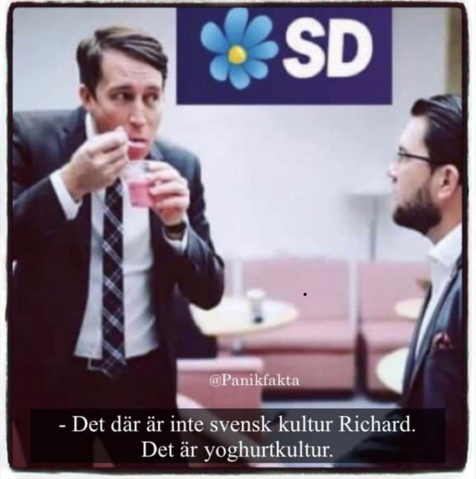 Två män i kostymer i en diskussion, den ena äter yoghurt, med en plakat för SD i bakgrunden.