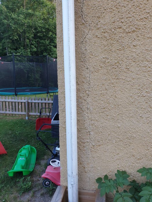 Vertikal spricka i putsad fasad på hus nära hörn och regnrör, med trädgårdsleksaker i förgrunden.