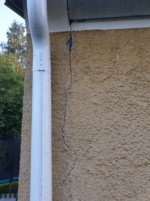Spricka i hustomtens puts från taknock ned mot grund intill stuprör, synlig skada på putsad yta.