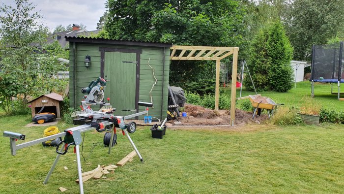 Nybyggd träpergola i trädgårdshörn med verktyg och material framför grönt förråd.