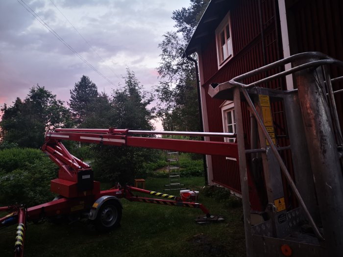 Skylift parkerad vid rödmålat hus i skymningsljus efter användning för renoveringsarbete.