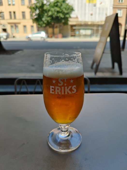 Ett glas öl av märket St. Eriks på ett bord med suddig stadsmiljö i bakgrunden.