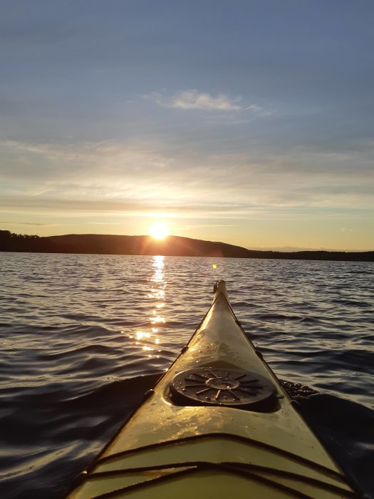 Vy från en kajak som glider på en stilla sjö vid solnedgång med solens reflexion på vattnet.