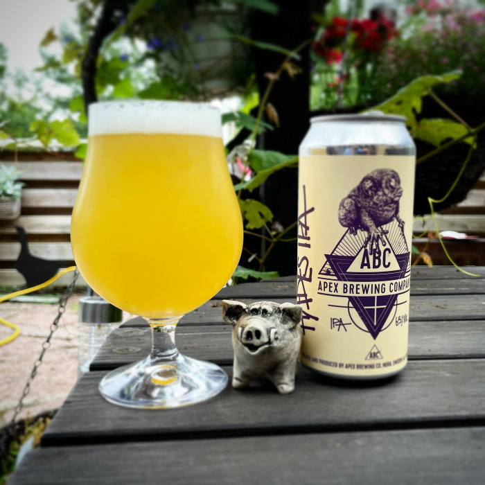 En grumlig öl i ett glas bredvid en burk från Apex Brewing och en liten grisfigur på ett trädäck.