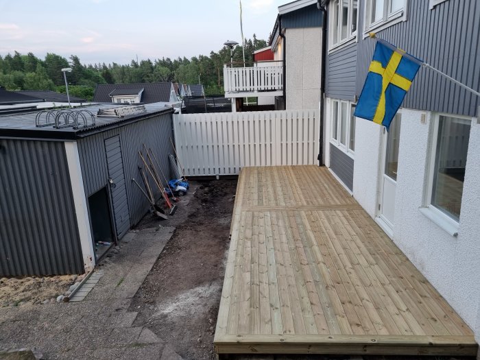 Nybyggd träaltan bredvid ett hus, med ett pågående markarbetsprojekt och en svensk flagga.