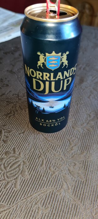 Öppnad ölburk med etiketten "Norrlands Djup" och en alkoholstyrka på 6,8% volym placerad på ett bord med mönstrad duk.