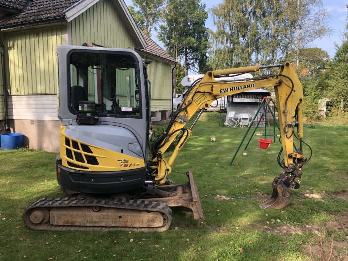 Gul och grå New Holland 2,7t minibagrare med grävskopa på en gräsmatta framför ett grönt hus.