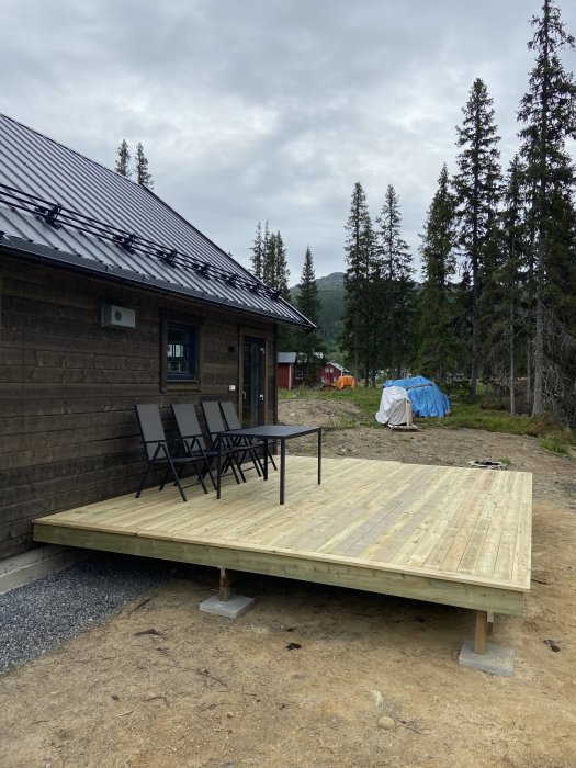 Nyligen byggd träaltan på betongplattor bredvid trähus med trädgårdsmöbler, omgiven av skog och berg.