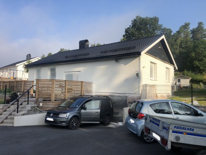 Nyrenoverat vitt hus med svart tak under solig himmel, bilar och släpvagn framför.