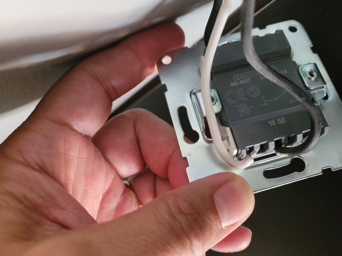 Hand som installerar en grå elektrisk strömbrytare med kopplingsanvisningar och kabelanslutningar synliga.