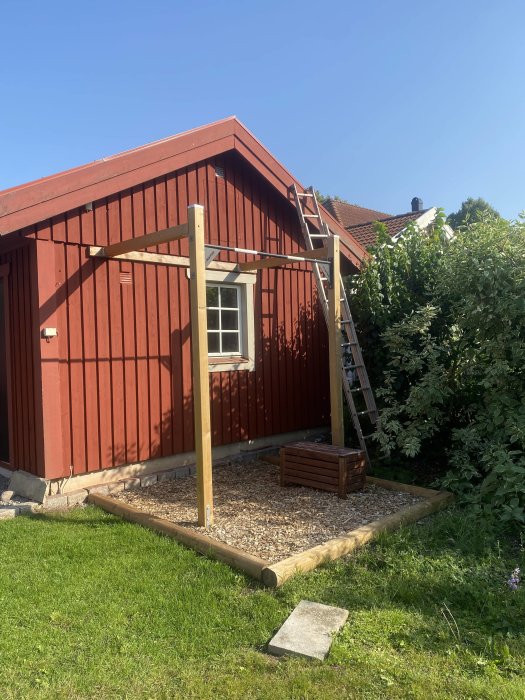 Träställning med stolpar och pull-up stång fastsatt vid röd garagewand, omgiven av gräs och buskage.