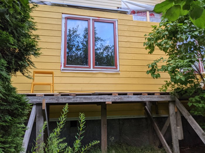 Gulpanelad husvägg med kvalitetsbrister och en röd fönsterram, förarbete för vidare renovering synligt.