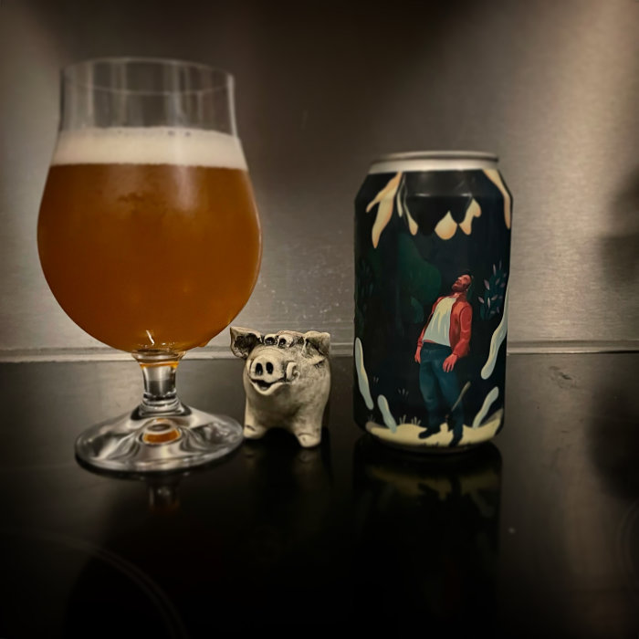 Ölglas fyllt med bärnstensfärgad dryck, konstnärligt ölburk med illustration, liten griskeramik till vänster.