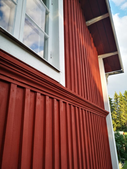 Nymålad röd locklistpanel på husfasad vid ett fönster med synliga hörn och detaljer.