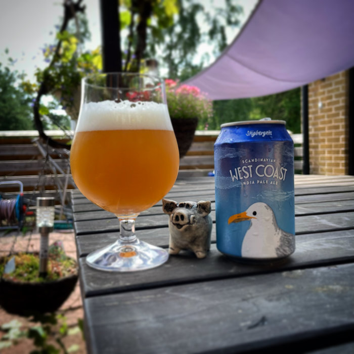 Ölglas med skummande öl och en burk IPA på ett utomhusbord, med en liten grisfigur och trädgård i bakgrunden.