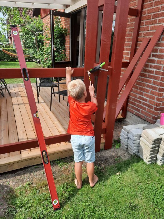 Barn hjälper till med byggarbete genom att mäta med vattenpass på en rödmålad träkonstruktion utomhus.